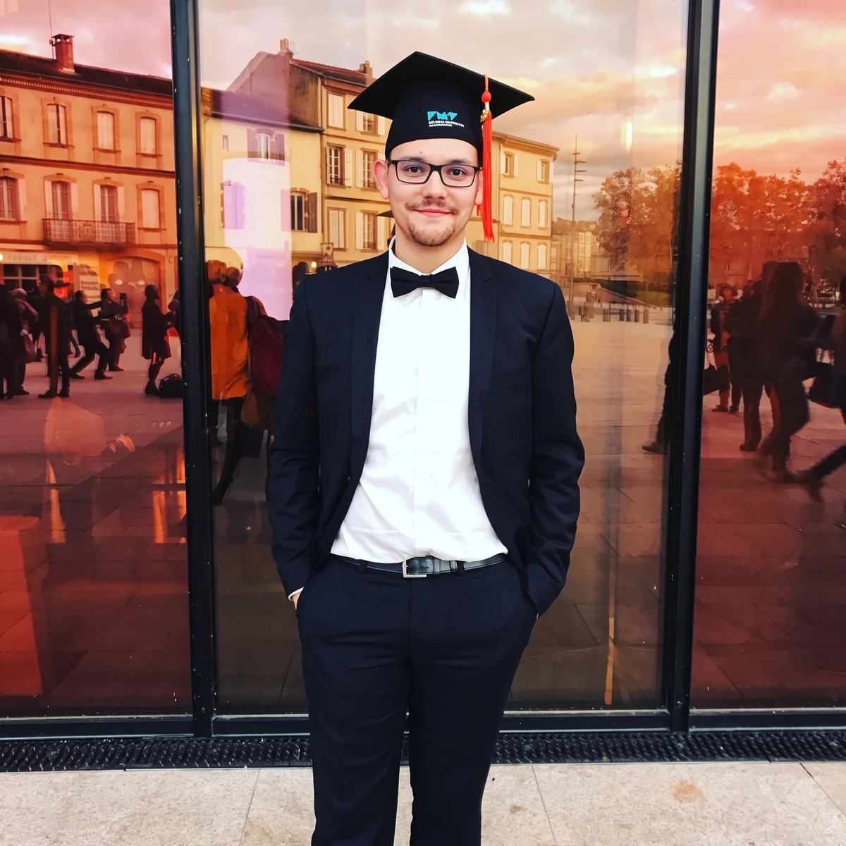 Alexis en costume, nœud papillon et toque de diplômé, devant une vitrine à Albi